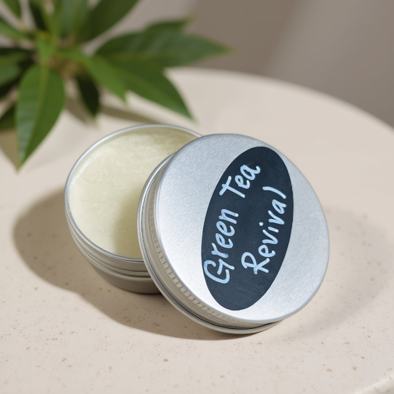 Tin of 'Green Tea Revival' cream on a light surface with green leaves in the background