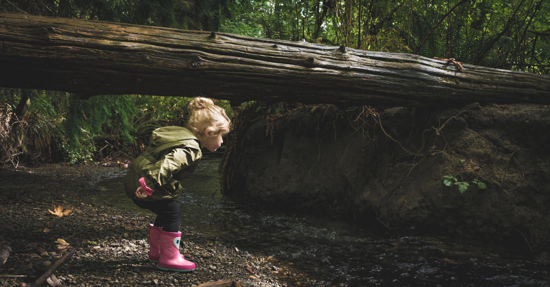 A child in pink rain boots bending over to explore a wooded stream